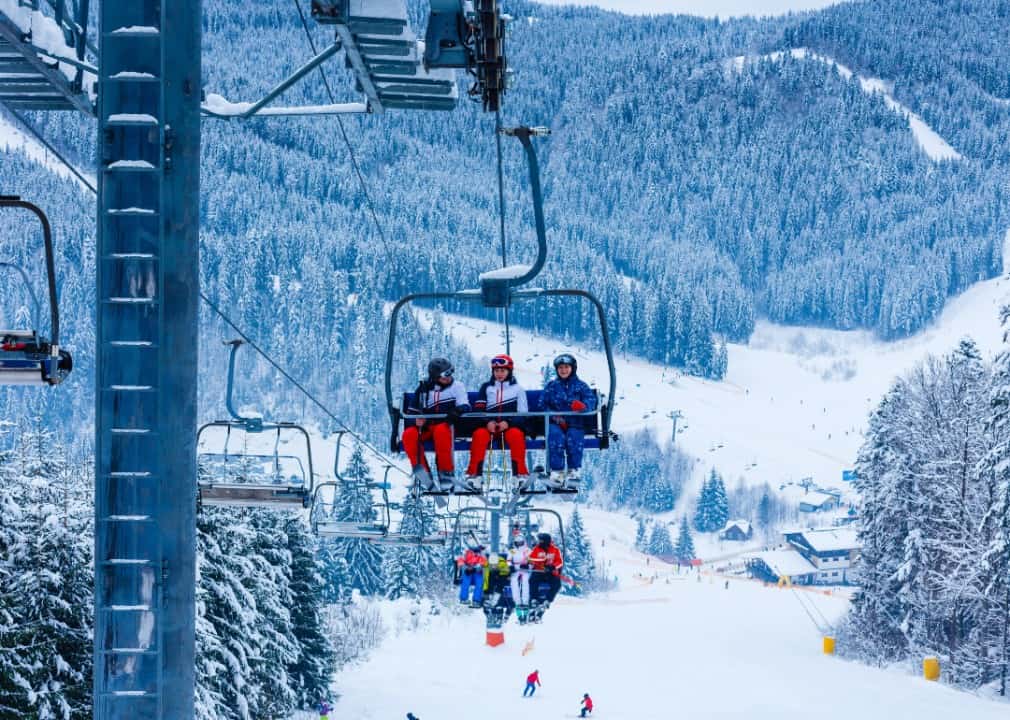 Ski lift with people going up the mountain and views of mountains covered in snow in the background. 