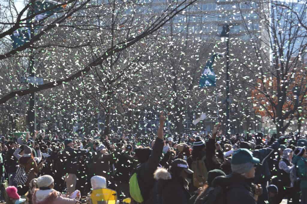 A packed crowd cheering during the Eagles Super Bowl Parade, with green and white confetti flying through the air.